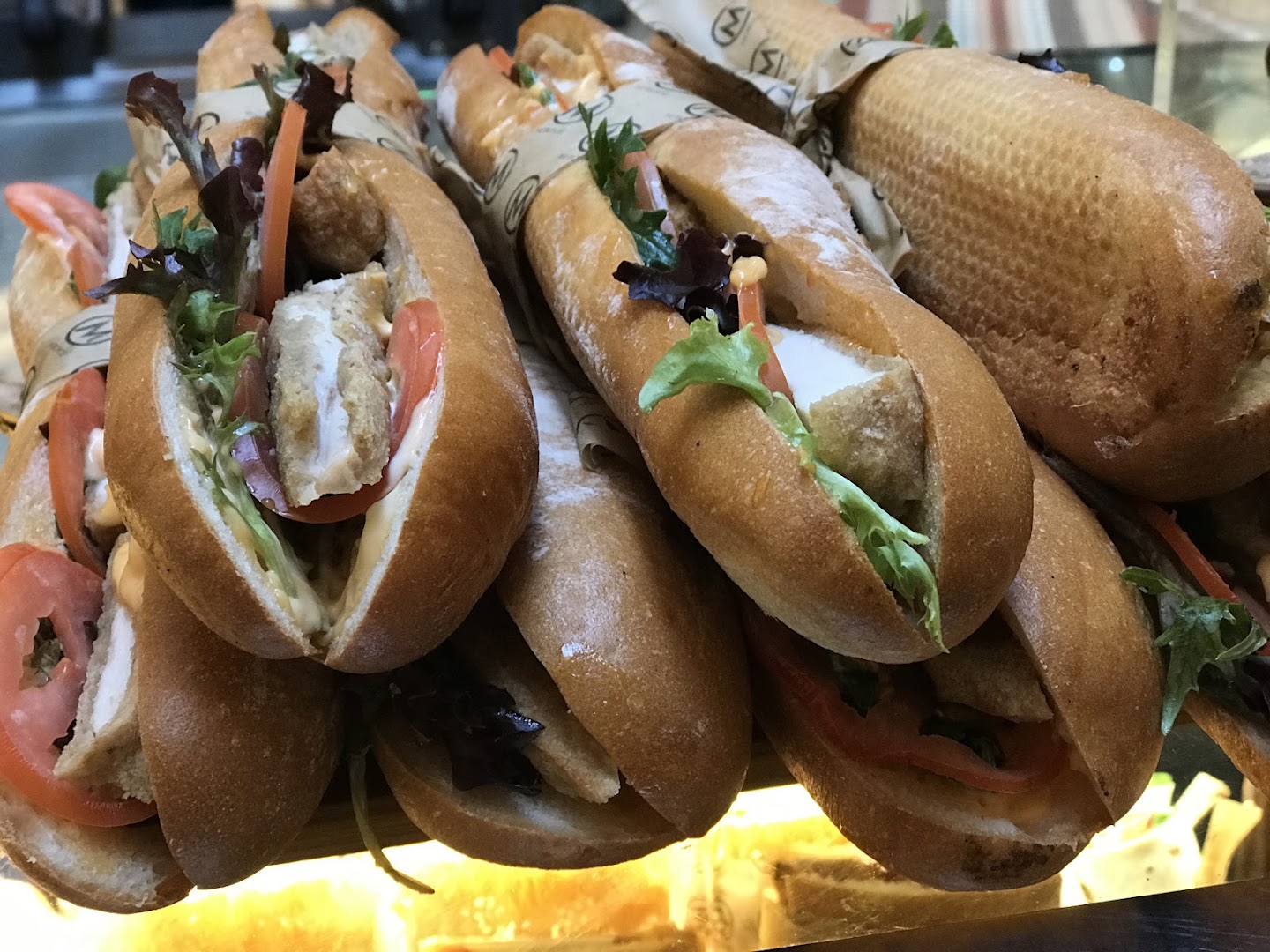 Filled baguette rolls stacked in a lit cabinet, each half wrapped in printed paper. Salad leaves, sliced tomato and crumbed fillings show at the open ends.