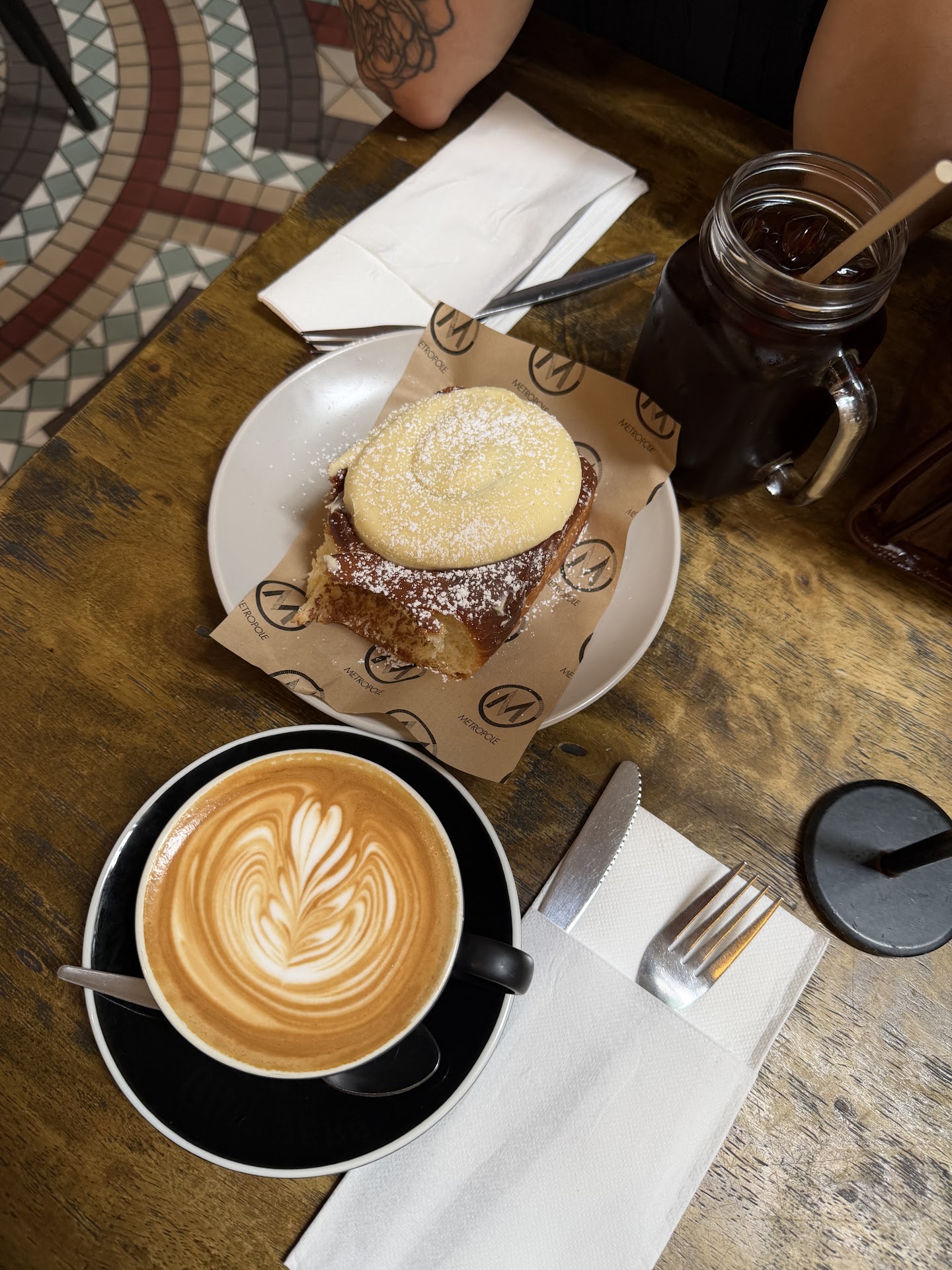 A table from above: a coiled bun with a pale set topping and a dusting of sugar on a plate lined with Metropole-printed paper, a coffee with poured latte art in a black cup, a jar of iced black coffee with a straw, cutlery on paper napkins, and the edge of the mosaic floor at the top of the frame.