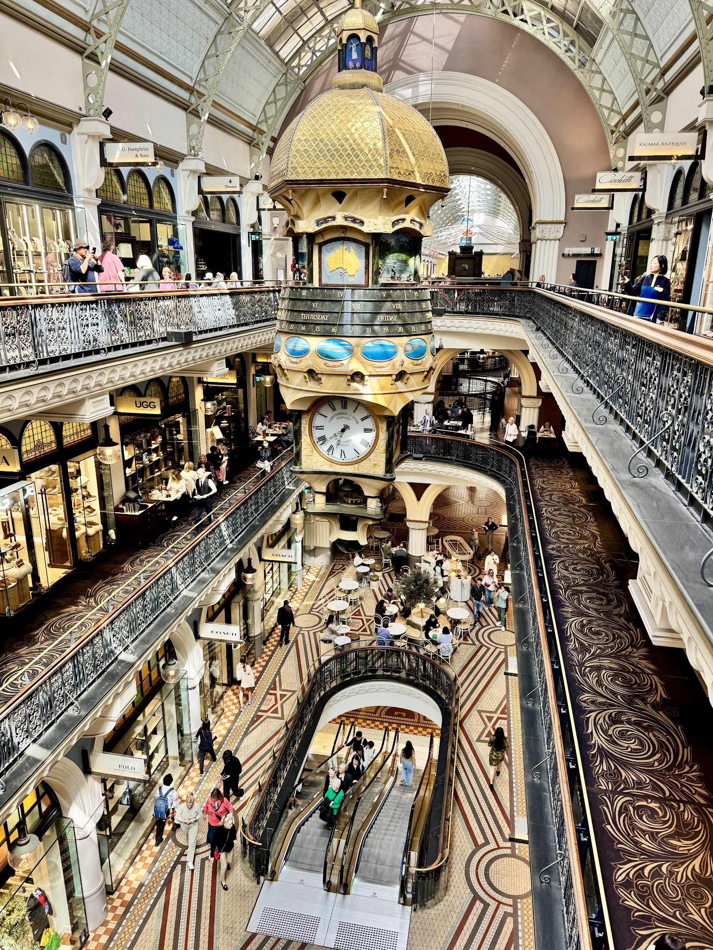 The Queen Victoria Building seen from an upper gallery: a large gilded hanging clock with a domed top and a white dial, wrought-iron balustrades on two levels, a glazed barrel roof, shopfronts, escalators, and a patterned tile floor with seating at the far end.