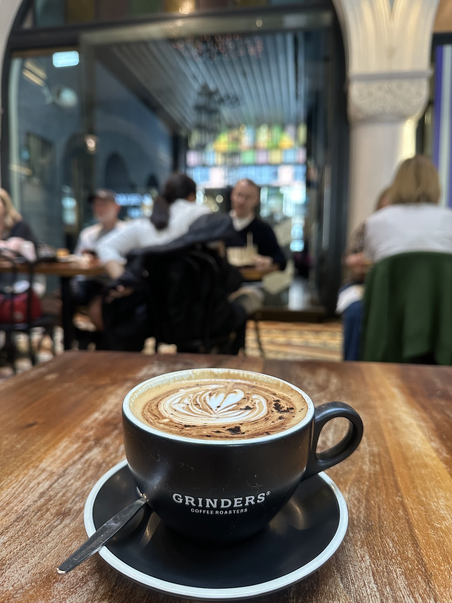 A cappuccino with poured latte art in a black cup and saucer on a worn wooden table, photographed low. Behind it, out of focus, people sit at tables under a tall arched opening with stained glass beyond.
