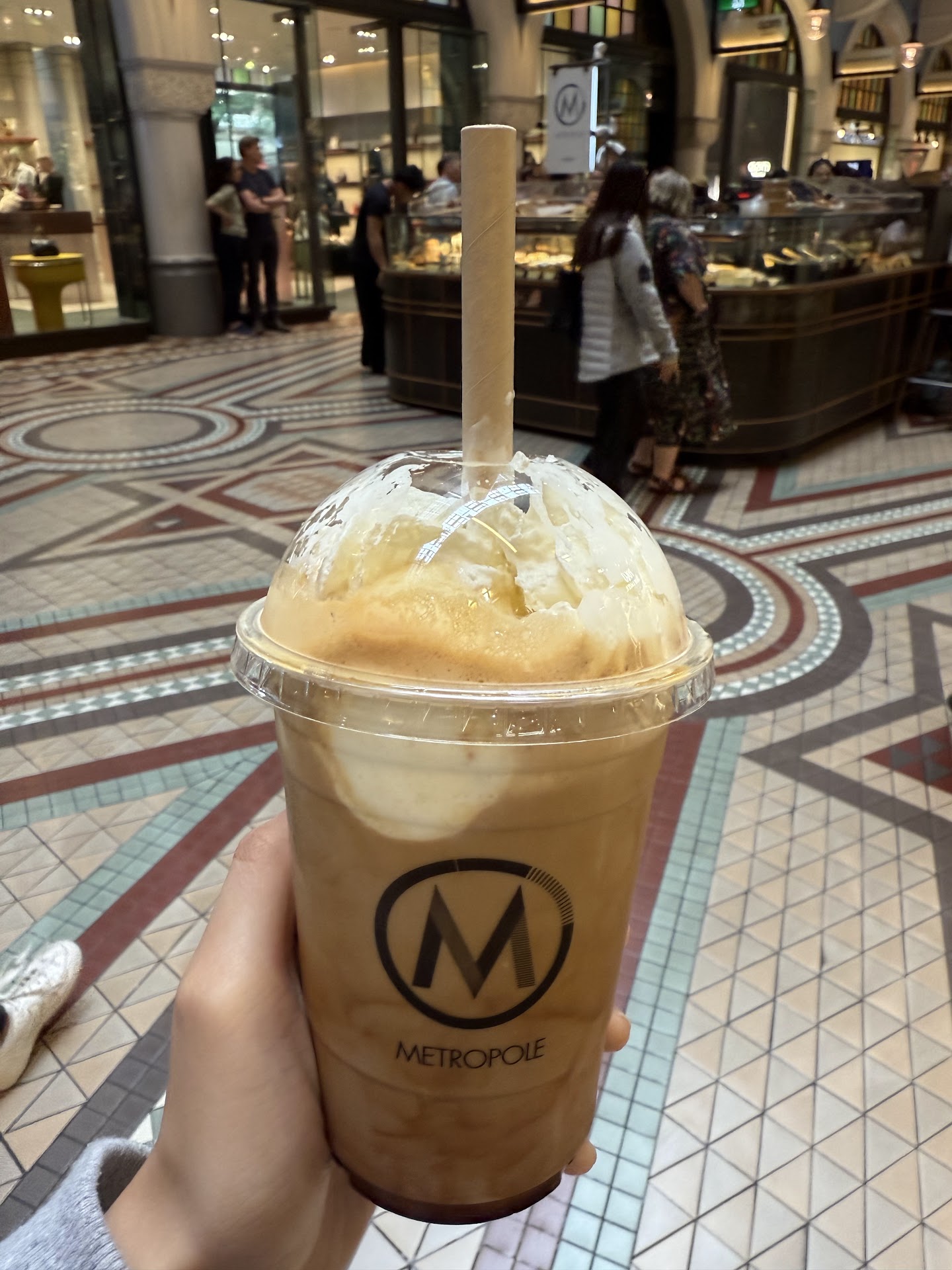A hand holds up a clear takeaway cup of iced coffee topped with cream, printed with the round Metropole M mark and a paper straw. Behind it the kiosk and its cabinet, people standing at it, and the geometric mosaic floor of the arcade.
