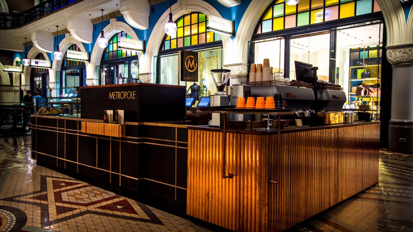 The Metropole kiosk on the ground floor of the Queen Victoria Building, empty and lit before opening: a long counter with a vertically slatted timber front and brass banding, METROPOLE lettering on the black service block, an espresso machine with stacked orange cups, a grinder, and a round M sign hanging behind it. Stone arches, stained glass and a patterned mosaic floor surround it.