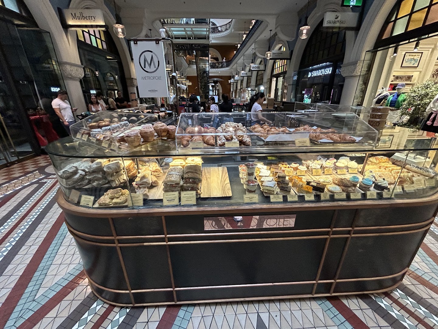 The kiosk in trade, seen across the mosaic floor: a long glass-topped cabinet of croissants, pastries, wrapped rolls and labelled slices, a dark curved base with brass banding and METROPOLE lettering, a hanging M sign, staff working behind, and shopfronts in the arcade beyond.