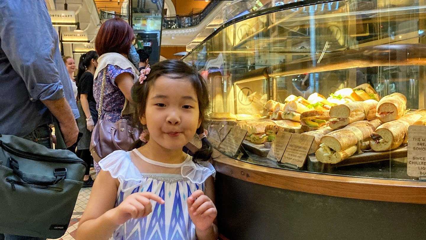 A child stands smiling beside the kiosk's curved glass cabinet, which is full of rolls on labelled boards. Adults queue behind, and the galleries of the Queen Victoria Building rise out of focus in the background.