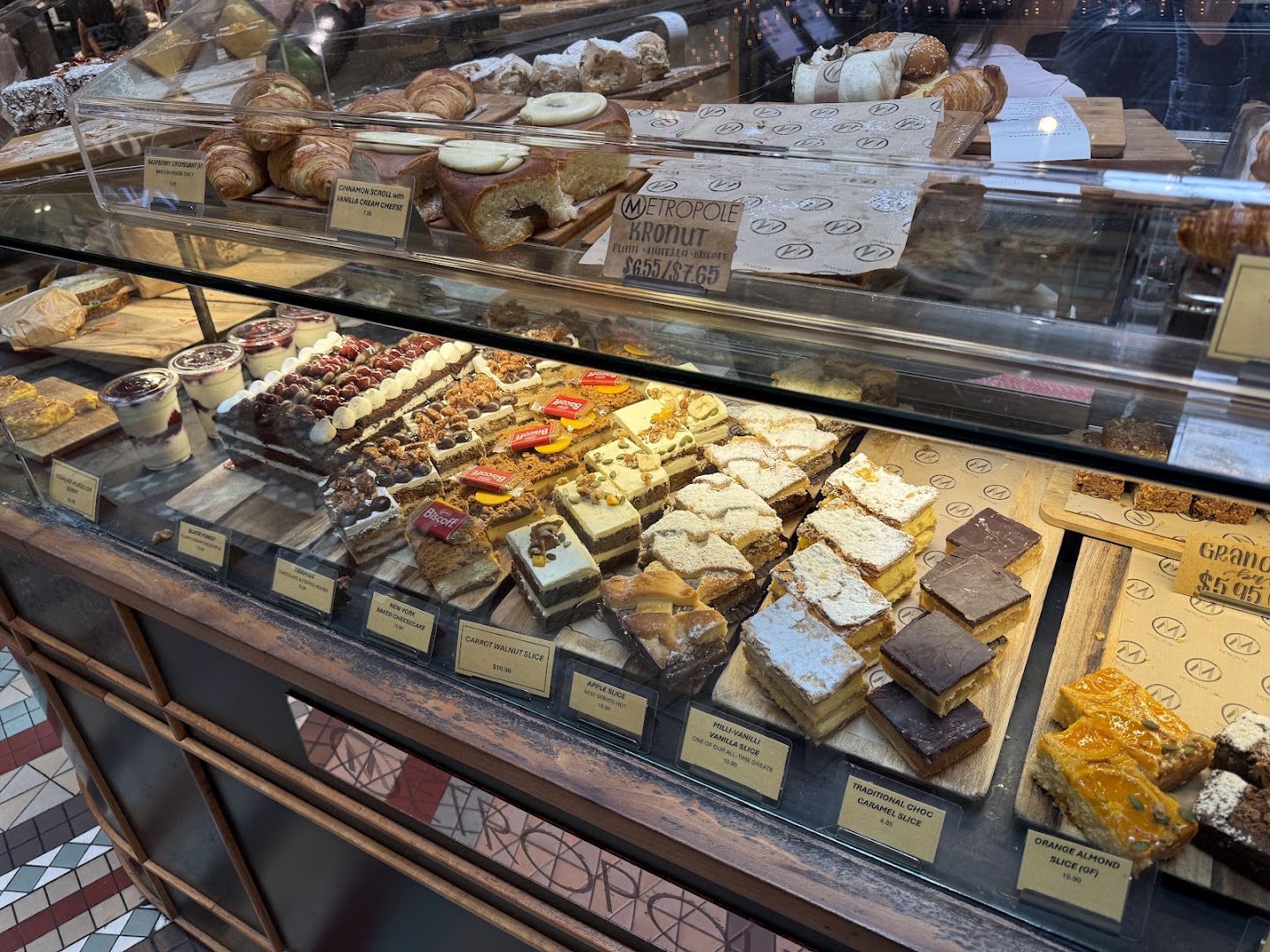 Close on the cabinet: two tiers of slices, tarts and layered cakes on paper-lined boards, each with a small hand-written ticket in front of it, croissants and pastries on the shelf above, and jars of layered dessert at the left end.